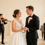 Quelles chansons choisir pour un mariage à l’image du couple et des temps forts ? Un couple de mariés souriant pendant une cérémonie laïque, avec un violoniste et un pianiste en arrière-plan dans une lumière douce.