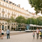 Quels quartiers ou communes choisir à Montpellier selon son budget d’achat ? Vue urbaine réaliste de Montpellier avec des immeubles anciens en pierre, un tramway moderne et des familles se promenant dans une place arborée sous une douce lumière méditerranéenne.